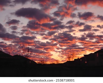 A vibrant sunset with dark clouds tinged in shades of orange and pink, filling the sky above silhouetted rooftops and leafless trees. - Powered by Shutterstock