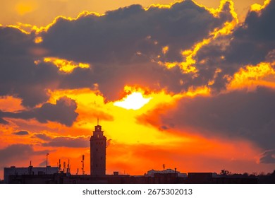 A vibrant sunset illuminates the sky, casting warm colors over a historic tower. The clouds create dramatic patterns as the sun descends, marking the end of a beautiful day.
