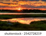 Vibrant sunset over a marsh in Acadia National Park, Maine, with the sky ablaze in orange and pink hues reflected on calm water, creating a peaceful and picturesque scene