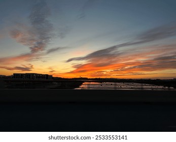 A vibrant sunset paints the sky with orange and pink hues, reflecting over calm water with silhouetted buildings in the distance. This serene urban scene captures the peaceful beauty of dusk - Powered by Shutterstock