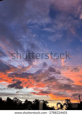 A vibrant sunset sky filled with dramatic purple, pink, and orange clouds above a quiet neighborhood. Silhouettes of trees and power lines frame the glowing horizon, creating a colorful evening atmosphere.