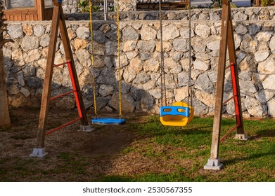 A vibrant swingset made of wood and metal stands in a lush grassy area, surrounded by rustic stone walls. The swings sway gently in the breeze, promising fun for children. - Powered by Shutterstock