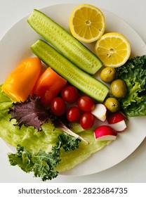 A vibrant top-down flat lay of assorted fresh vegetables arranged on a white plate, including sliced cucumbers, cherry tomatoes, orange and red bell peppers, green olives, radish wedges, lemon halves, and various lettuce leaves. The clean white background highlights the natural colors and freshness of the produce. Ideal for concepts related to healthy eating, fresh produce, salads, nutrition, and clean living.