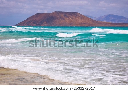 Vibrant turquoise waves roll gently toward the shore of Fuerteventura, with Lobos Island rising in the distance under a cloudy sky. A stunning seascape capturing the raw beauty of the Canary Islands