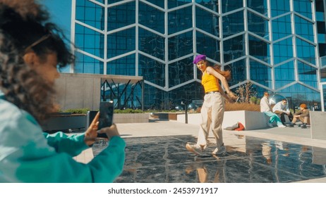 Vibrant Urban Dance Scene With A Young Woman In A Purple Beanie And Yellow Top Performing A Dynamic Dance Move, Captured By A Friend On A Smartphone, Against A Modern Glass Building Background. - Powered by Shutterstock