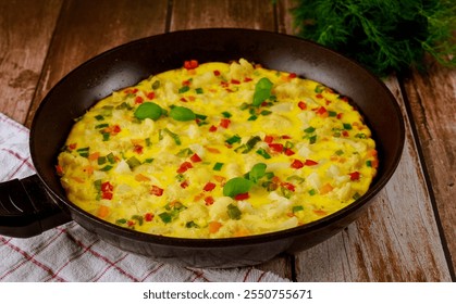 Vibrant vegetable omelette being prepared in black frying pan, featuring colorful bell peppers, herbs, and onions on rustic wooden table with cloth.