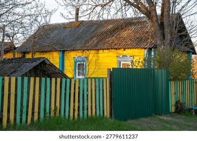 Vibrant yellow timber cottage features blue window frames and weathered slate roof standing behind green and yellow picket fence in sunny rural courtyard.