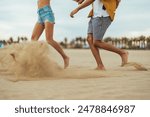 A vibrant young Hispanic couple enjoys playfully kicking up sand as they engage in leisure activities on a scenic beach, showcasing their casual summer attire.