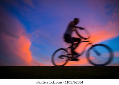 Vibrantly Colored View Of Sunset Clouds Glowing Behind The Motion Blur Silhouette Of A Man Riding A Beach Cruiser Bike Across The Horizon