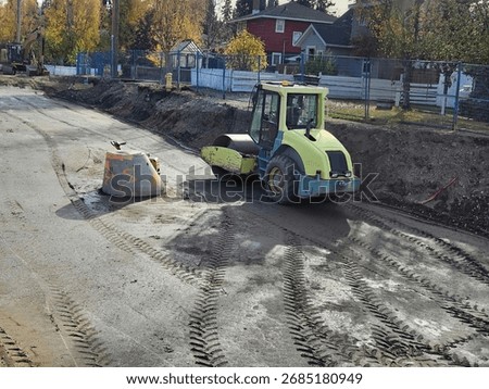 Vibratory drum roller compacts urban subgrade around precast concrete manhole section during road reconstruction.
