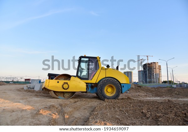 Vibro Roller Soil Compactor leveling ground at
construction site. Vibration single-cylinder road roller on
construction road. Road work for new asphalt laying. Tower cranes
build high-rise buildings