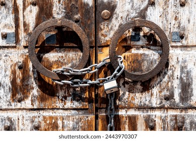 Vicenza, Venetien - Italy - 06-12-2021: A shiny chain and silver padlock secure a weathered wooden door with rusty iron rings, combining old and modern