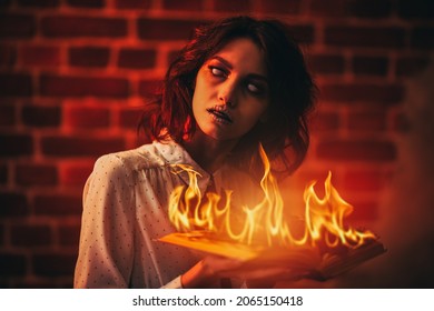 Vicious Ghost Girl With Faded Eyes Stands With A Burning Book In Her Hands In A Gloomy Room Lit By Sinister Red Light. Horror Scene. Halloween.