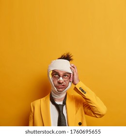 Victim Of Accident Has Severe Head Trauma, Stands Wrapped In Bandage, Lost Memory, Concentrated Above, Has Bruises And Adhesive Plaster On Nose, Dressed Formally, Isolated On Yellow Background