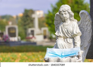 Victim Of Coronavirus Covid 19 Conceptual Photo With Sculpture Of A Little Angel Holding A Protective Medical Mask On A Tomb In Cemetery. Selective Focus