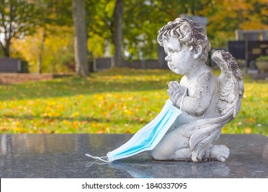 Victim Of Coronavirus Covid 19 Conceptual Photo With Sculpture Of A Little Angel Holding A Protective Medical Mask On A Tomb In Cemetery. Selective Focus