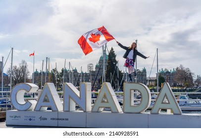 Victoria, British Columbia, Canada - March 19 2022: Girl holds windblown Canada flag while standing on a Canada sign in front of the BC legislature in preparation for Canada Day and Freedom rallies