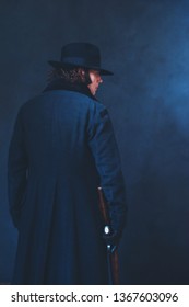 Victorian man in long black coat and hat holding rifle. Rear view.