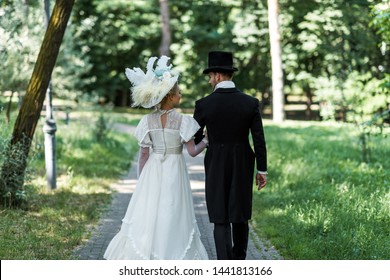 victorian man and woman in hats walking outside near green trees 