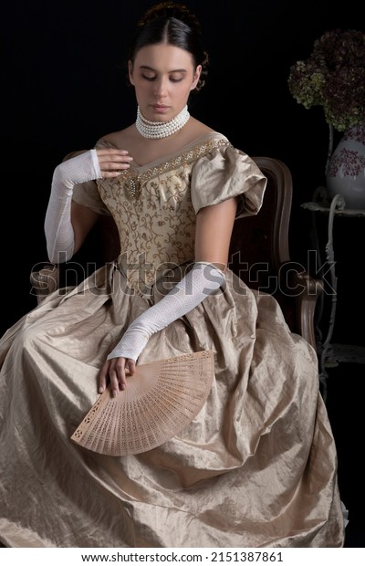 A Victorian woman in a gold ball\
dress sitting on an ornate chair against a dark\
backdrop