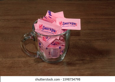 Victorville, CA / USA – December 18, 2019: Pink Packets Of The Artificial Sweetener Band Of Sweet ’N Low In A Glass Coffee Cup On A Brown Table.