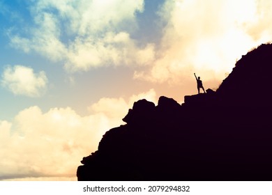 Victory On Top Of A Mountain. Female Hiker Silhouette Standing On A Rocky Mountain Edge With Fist Up To The Sky.