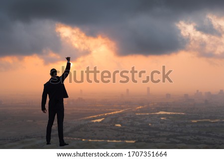 Victory and success concept! Strong confident businessman celebrating on top of skyscraper overlooking against a city sunrise background. 