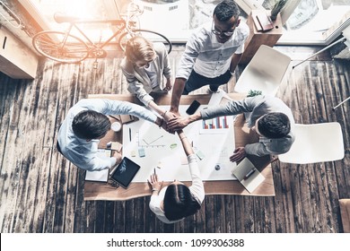 Victory!  Top view of young modern people in smart casual wear holding hands on top of one another in a symbol of unity while working in the creative office