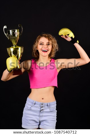 Victory, winner and success concept - excited and joyful fighter boxer girl wearing boxing gloves celebrates victory. Smile woman in sportswear with boxing gloves holds gold winner trophy. Boxer woman