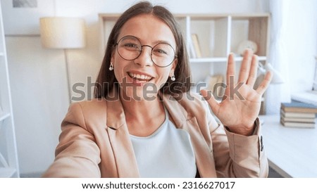Video blog. Influencer streaming. Beautiful happy expressive young woman with big smile in joyful mood in glasses waving hand to camera for warm greeting in white room.