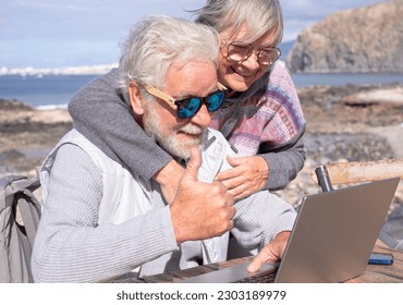 Video call concept. Cheerful bonding senior family couple using laptop in video chat enjoying a sunny day outdoor at the beach. Elderly people in vacation or retirement at sea enjoying tech and social - Powered by Shutterstock