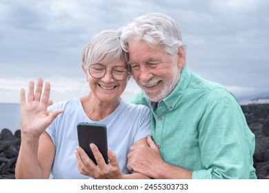 Video call concept. Happy senior couple sitting outdoors by the sea waving hand in video connection via mobile phone webcam. Cloudy sky in the background and horizon over the water - Powered by Shutterstock