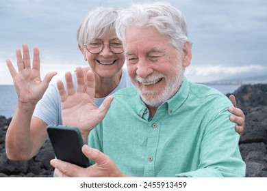 Video call concept. Happy senior couple sitting outdoors by the sea waving hand in video connection via mobile phone webcam. Cloudy sky in the background and horizon over the water - Powered by Shutterstock