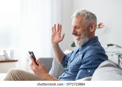 Video call concept. Hilarious grey-haired middle aged hipster man in casual shirt using smartphone for virtual meeting sitting on the sofa at home, mature guy enjoys online chatting, waving hi - Powered by Shutterstock