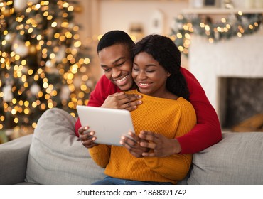 Video call on eve of Christmas. Family happy millennial african american husband and wife look at tablet and congratulate friends in cozy living room interior with Xmas tree and glowing garlands - Powered by Shutterstock