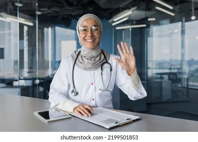 Video call online consultation, woman in hijab Muslim doctor looking at camera and waving greeting gesture, female doctor consulting patients remotely, working in modern clinic office - Powered by Shutterstock