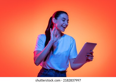 Video Call. Portrait Of Young Emotional Girl, Student In White T-shirt Isolated On Orange Color Background In Neon Light. Concept Of Beauty, Art, Fashion, Education