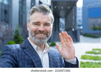 Video Call Of Senior Mature Businessman, Gray Haired Man Looking Phone Camera And Smiling, Waving Hand Gesture Of Greeting, Investor Outside Office Building Talking Remotely With Friends Colleagues