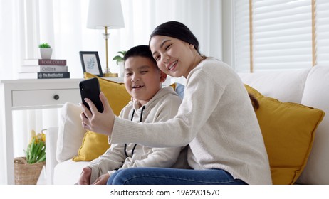 Video Call Teleconference Asian Family With Young Mother And Son Using Mobile Phone Internet Wifi Remotely Chat At Home Living Room Feeling Happy In Self Isolate Quarantine From Coronavirus Covid.