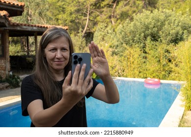 Video Calling, Portrait Of Happy Blonde Woman Video Calling. Making FaceTime With Smartphone At Vacation. Work From Holiday, Standing Before Swimming Pool, Using Meeting Online Application, Wave Hand.