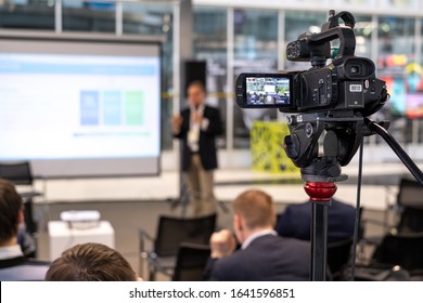 Video camera on tripod recording presentation of male coach standing on stage with display in front of audience