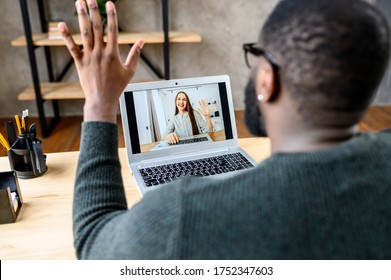 Video chat with employee. Cheerful African-American guy in glasses waving hello to female coworker on the laptop screen. Back view black young man using laptop for video connection