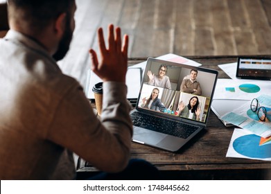 Video Communication. Back View Of A Young Man Who Communicates By Video Communication With His Employees, He Greets Them Waving His Right Hand. Distant Communication. Work From Home