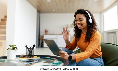 Video Conference. Portrait of cheerful African American woman in headset having web call using tablet pc sitting on pouf chair, talking waving hand at webcam in home office, free copy space, banner - Powered by Shutterstock