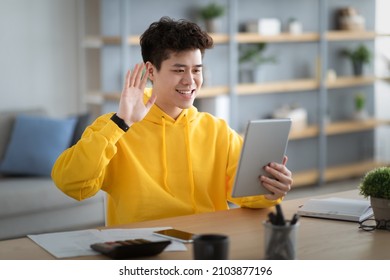 Video Conference. Portrait Of Cheerful Asian Man In Yellow Hoodie Having Web Call Using Tablet Pc Sitting At Desk, Talking Waving Hand At Webcam In Home Office, Free Copy Space, Blurred Background