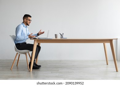 Video Conference. Profile Side View Portrait Of Middle Eastern Businessman In Glasses Having Web Call Using Laptop Sitting At Desk At Workplace, Talking And Gesturing At Webcam, Free Copy Space Banner