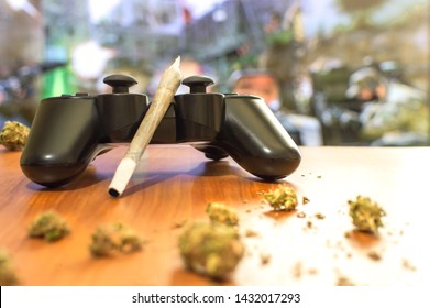 Video games and cannabis. Video game controller, big joint and grinder with marijuana buds on wooden table. Blur background of tv with video game.
