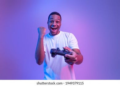 Video Gaming Concept. Euphoric African American Guy With Joystick Celebrating Game Win, Cheerful Black Man Raising Fist And Exclaiming With Excitement, Standing In Neon Light Over Purple Background