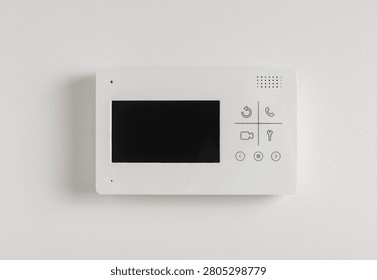 A video intercom panel with a small screen and buttons is mounted on a light textured wall. The doorphone device allows residents to view and communicate with visitors before granting access. A
