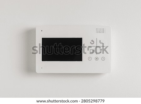 A video intercom panel with a small screen and buttons is mounted on a light textured wall. The doorphone device allows residents to view and communicate with visitors before granting access. A